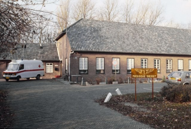 Ambulanz vor einem Backsteingebäude mit Fenstern, einem Schild, Pflanzen, Bäumen und einem klaren blauen Himmel im Hintergrund geparkt.