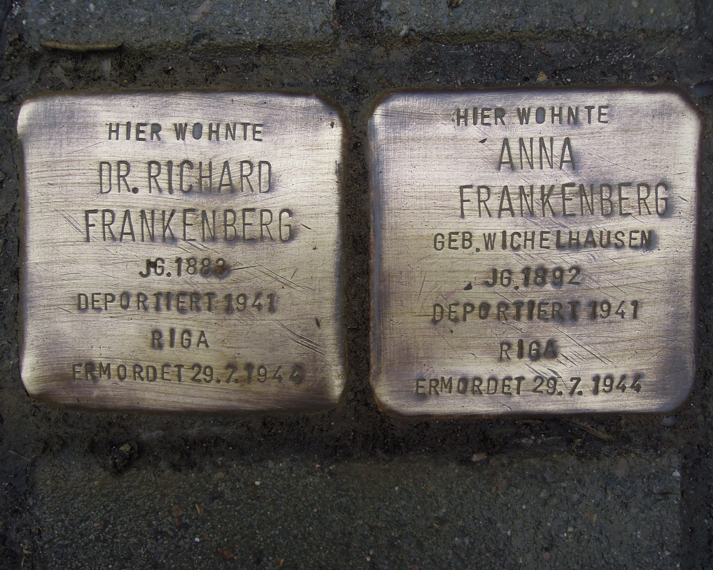 Zwei Messingplaketten vor einem Gebäude auf dem Boden, mit Text beschriftet, der Holocaust-Opfer gedenkt.