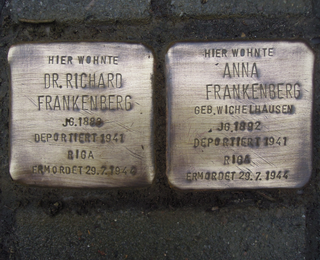 Zwei Messingplaketten vor einem Gebäude auf dem Boden, mit Text beschriftet, der Holocaust-Opfer gedenkt.