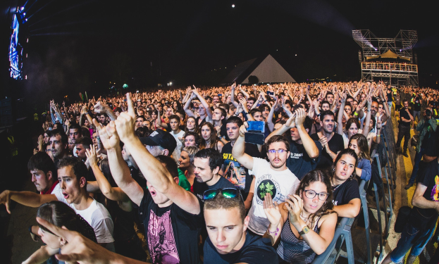 Eine große Menge bei einem Musikfestival mit erhobenen Händen, einige sitzen auf Stühlen und andere stehen, begrenzt von einem Metallzaun auf der rechten Seite, mit Gebäuden und Lichtern im Hintergrund unter einem dunklen Himmel, mit einem Mann und einer Frau im Vordergrund.