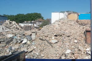 Ein großer Schutthaufen inmitten einer Baustelle, umgeben von Gebäuden, Bäumen und einem klaren blauen Himmel, mit einer Abrissmaschine am unteren Bildrand.