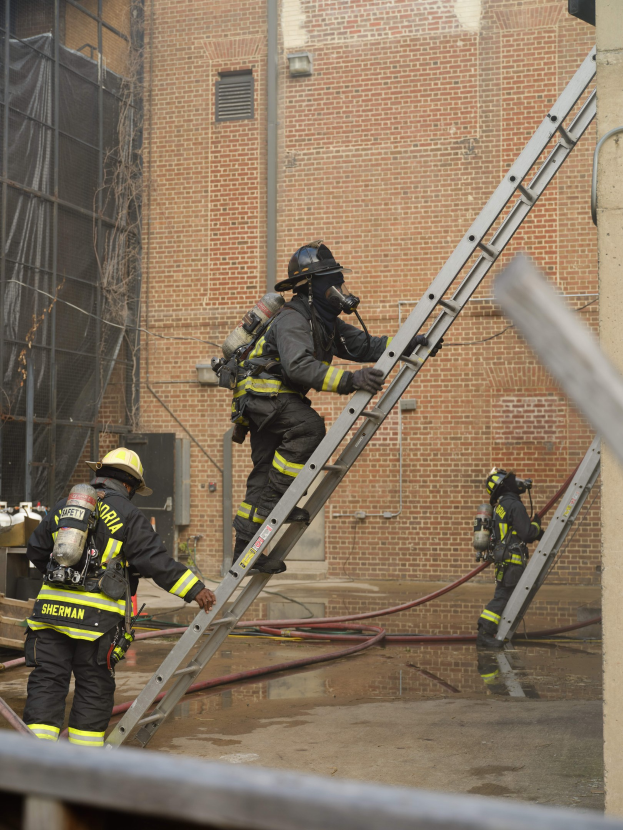 Feuerwehrleute in Helmen und Ausrüstung klettern eine Leiter vor einem Backsteingebäude mit Rohren auf dem Boden und einem Metallstab an der Basis hoch, mit einem anderen Gebäude und einem Netz im Hintergrund.