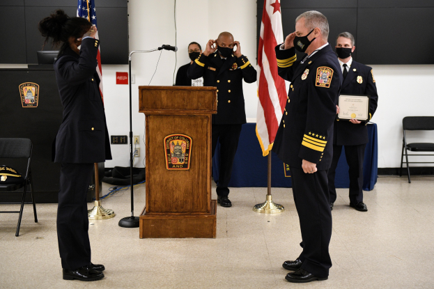 Gruppe uniformierter Personen in Masken vor einem Rednerpult mit Mikrofon, umgeben von Fahnen, Stühlen, Tischen und einer Wand, mit einer Person, die ein Zertifikat während einer Vereidigung eines neuen Feuerwehrchefs hält.