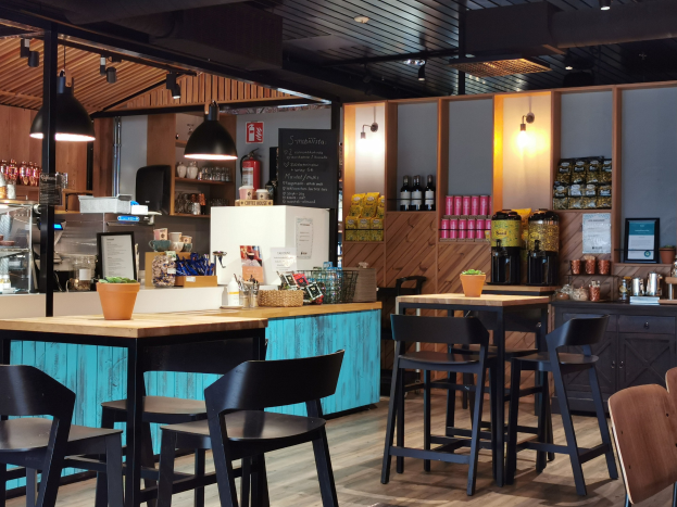 Innenansicht eines Restaurants mit Holztischen, Stühlen, einer Topfpflanze auf einem Tisch, einer Schiefertafel, einem Kühlschrank, einem Feuerlöscher, Regalen mit Flaschen, Schränken mit Gegenständen, einem Rahmenfoto an der Wand und Deckenleuchten.