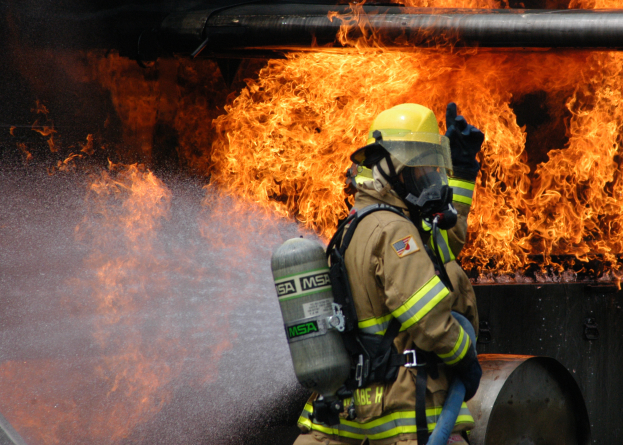 Feuerwehrmann in Schutzausrüstung nutzt einen Schlauch, um Wasser auf ein Feuer zu sprühen, mit einem brennenden Rohr im Hintergrund.