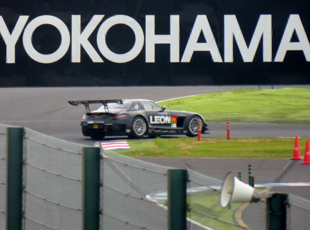 Schwarzer Sportwagen auf einer Rennstrecke umgeben von Verkehrskegeln, mit einem "Yokohama"-Schild im Hintergrund, Gras, einem Zaun und einem Lautsprecher im Vordergrund.