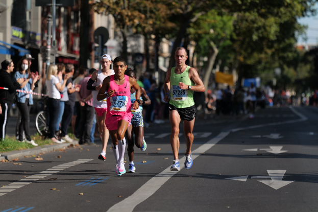 Gruppe von Läufern bei einem Stadtmarathon, mit Zuschauern links, unscharfer Stadtpanorama, Bäumen und einem Fahrrad im Hintergrund.