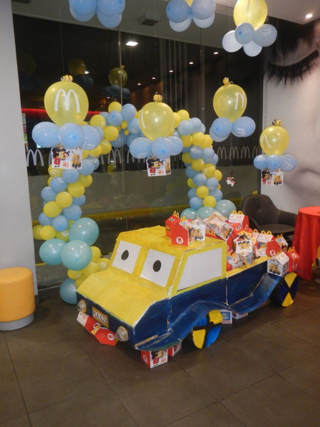 Eine McDonald's-Geburtstagsfeier mit einem gelben und blauen Lastwagen, der mit Ballons und anderen Dekorationen geschmückt ist, einem rot eingedeckten Tisch mit Gegenständen, Hockern, einer Glaswand und Deckenlampen in einer festlichen Atmosphäre.