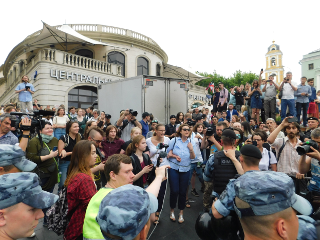 Eine große Gruppe von Menschen protestiert vor einem Gebäude, einige halten Kameras und Handys, mit einem geparkten Fahrzeug, Bäumen und einem klaren blauen Himmel im Hintergrund.