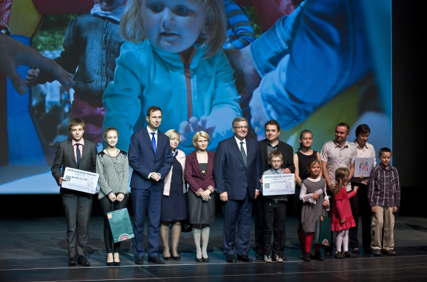 Eine Gruppe von Menschen auf einer Bühne mit Taschen und Urkunden, im Hintergrund ein Bildschirm mit einem Bild eines Kindes und anderer Personen.