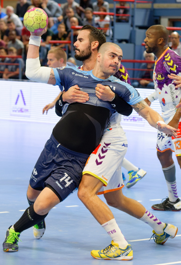 Männer beim Handballspielen in einer Stadion, einer hält den Ball, mit Zuschauern im Hintergrund.