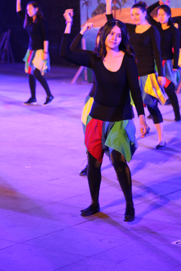 Eine Gruppe junger Frauen, die auf einer Bühne tanzen, mit einem Bild an der Wand im Hintergrund, alle tragen Schuhe.