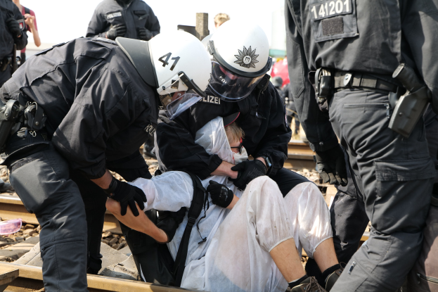 Eine Gruppe von Polizisten in Einsatzausrüstung verhaftet einen Mann in einem weißen Kleid auf einem Gleis, während andere im Hintergrund stehen.