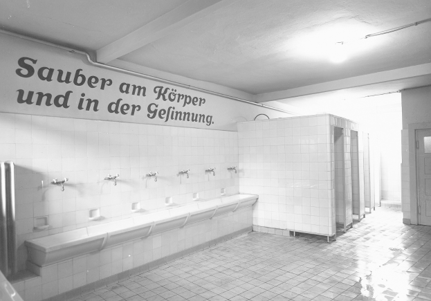 Schwarz-weiß-Foto einer öffentlichen Toilette mit Fliesenboden, einer Wand mit Text, mehreren Wasserhähnen, einer Tür und einer Deckenlampe.