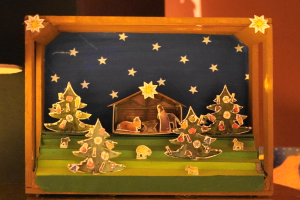 Eine hölzerne Krippe in einer Schachtel auf einem Tisch, mit einer Lampe links und einer Wand im Hintergrund, geschmückt mit Weihnachtsbäumen, Tieren und einem Haus.