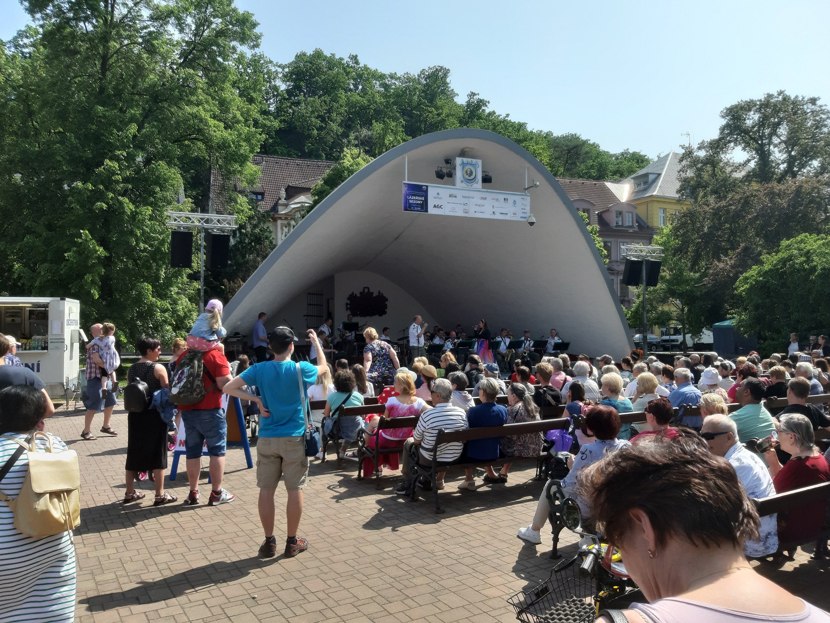 Menschenmenge auf Bänken vor einer Bühne mit stehenden Zuschauern, einem Kameramann, einem Mikrofonständer, einer Fahne, einem Metallrahmen, Bäumen, Gebäuden und einem bewölkten Himmel bei einem Sommerkonzert im Park.