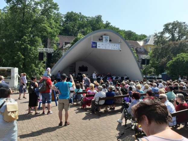 Menschenmenge auf Bänken vor einer Bühne mit stehenden Zuschauern, einem Kameramann, einem Mikrofonständer, einer Fahne, einem Metallrahmen, Bäumen, Gebäuden und einem bewölkten Himmel bei einem Sommerkonzert im Park.