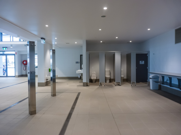 Große Öffentliches Klo mit mehreren Toiletten, tragenden Säulen, einem Mülleimer, einer Tür, einem Schild, einem angebrachten Feuerlöscher und Deckenleuchten auf dem Dach.