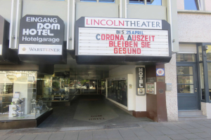 Das Lincoln Theater in Berlin, Deutschland, ein Gebäude mit Glasfenstern und -türen und einer Tafel mit Text darauf, zeigt verschiedene Objekte im Inneren und vermittelt den Eindruck einer pulsierenden Stadtlandschaft.
