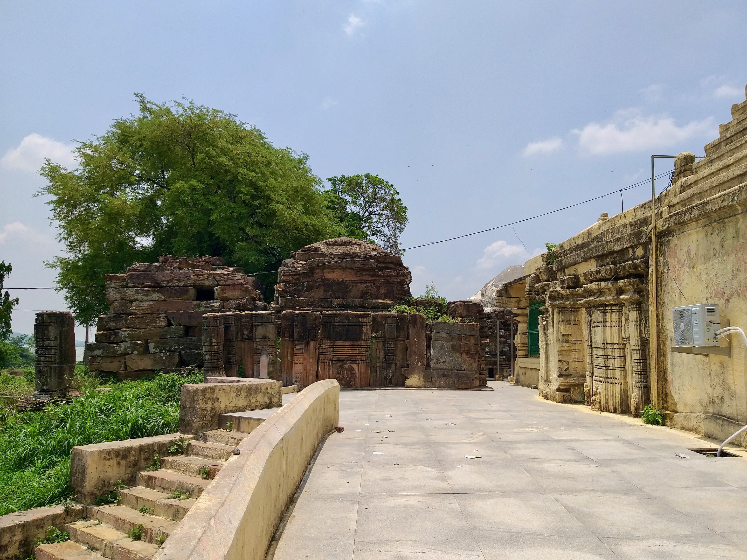 Ruinen eines Tempels mit alten Mauern, Treppen, Geländern, Pflanzen und Bäumen entlang einer Schotterstraße unter einem bewölkten Himmel.