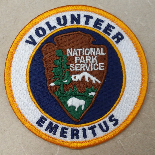Runder Aufnäher mit blauem Rand und weißem Hintergrund, mit schwarzem Text "Volunteer National Park Service Emeritus" in der Mitte eines gelben Kreises.