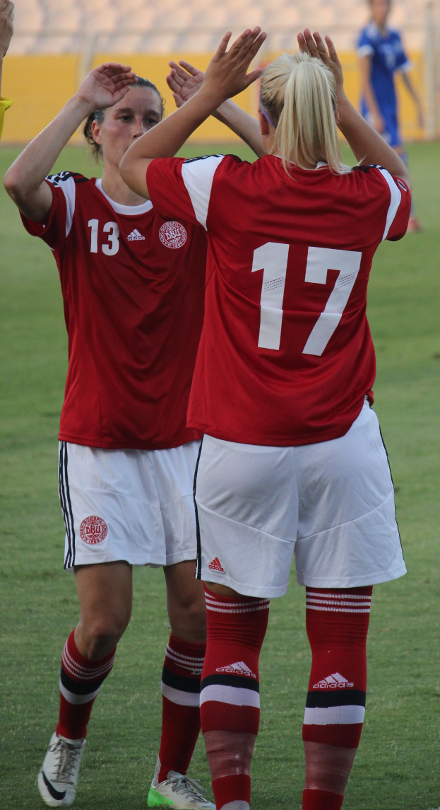 Zwei Frauen in roten T-Shirts und weißen Shorts stehen auf einem Fussballfeld und klatschen in die Hände, mit einer Person in einem blauen Kleid, einer gelben Tafel und einigen Stühlen im Hintergrund.