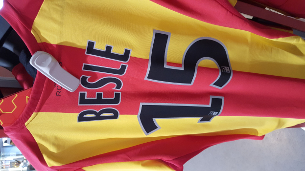 Roter und gelber Fußballtrikot mit aufgedrucktem Text und Nummern an einem Ladengestell hängend, mit anderen Gegenständen und dem Hintergrund im Hintergrund.