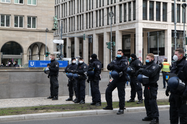 Polizeibeamte in schwarzen Uniformen und Masken stehen vor einem Gebäude mit Glasfenstern und Säulen in Berlin, Deutschland, einige halten Helme, mit Laternenmasten, Verkehrsampeln, Texttafeln, einer Statue und anderen Menschen in der Szene.
