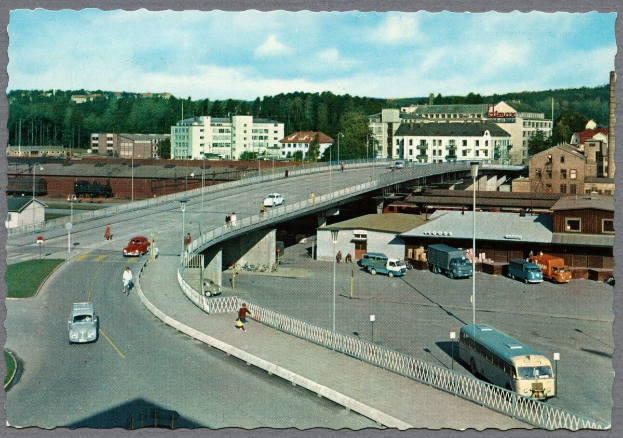 Altes Schwarz-Weiß-Foto einer Stadtstraße mit Autos, Bussen, Fußgängern auf einer Brücke, Laternen, Gebäuden, Bäumen und einem bewölkten Himmel.