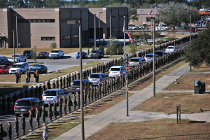 Eine große Gruppe von Menschen in militärischer Kleidung mit Gewehren steht vor einem Gebäude während eines Umzugs, mit Fahrzeugen, Laternenmasten, Fahnen, Bäumen und Gras im Vordergrund und Gebäuden unter einem klaren blauen Himmel im Hintergrund.