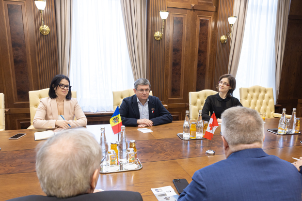 Eine Gruppe von Menschen sitzt um einen Holztisch in einem formellen Setting und diskutiert mit EU-bezogenen Dokumenten und Gegenständen, darunter Flaggen, Flaschen, Gläser, Papiere und Mobiltelefone.