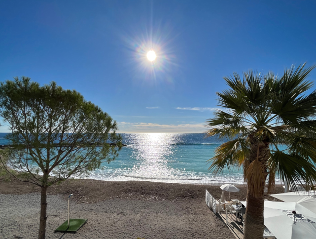 Ein sonniger Strand mit Palmen, Sonnenschirmen und üppiger Vegetation, vor einem strahlend blauen Himmel an der französischen Riviera.