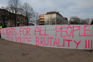 Eine Gruppe von Menschen, die auf dem Boden steht und ein Banner hält, auf dem 'Rechte für alle Menschen Stoppt Polizeigewalt' steht, mit einem Straßenpfahl, einem Schild, Bäumen, Gebäuden mit Fenstern und einem bewölkten Himmel im Hintergrund.