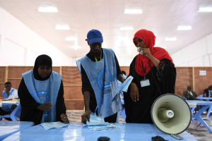 Eine Gruppe von Menschen steht um einen Tisch mit einem Megafon, Papieren und anderen Gegenständen an einer Wahllokal, während andere im Hintergrund sitzen und stehen unter beleuchteter Decke.