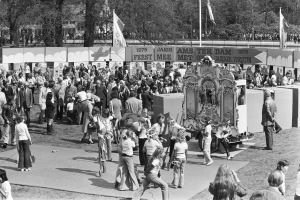 Schwarzes und weißes Bild einer Menge, die sich um einen mit Flaggen und Bannern geschmückten Karnevalswagen in einem Park versammelt hat, mit Bäumen und Gebäuden im Hintergrund.
