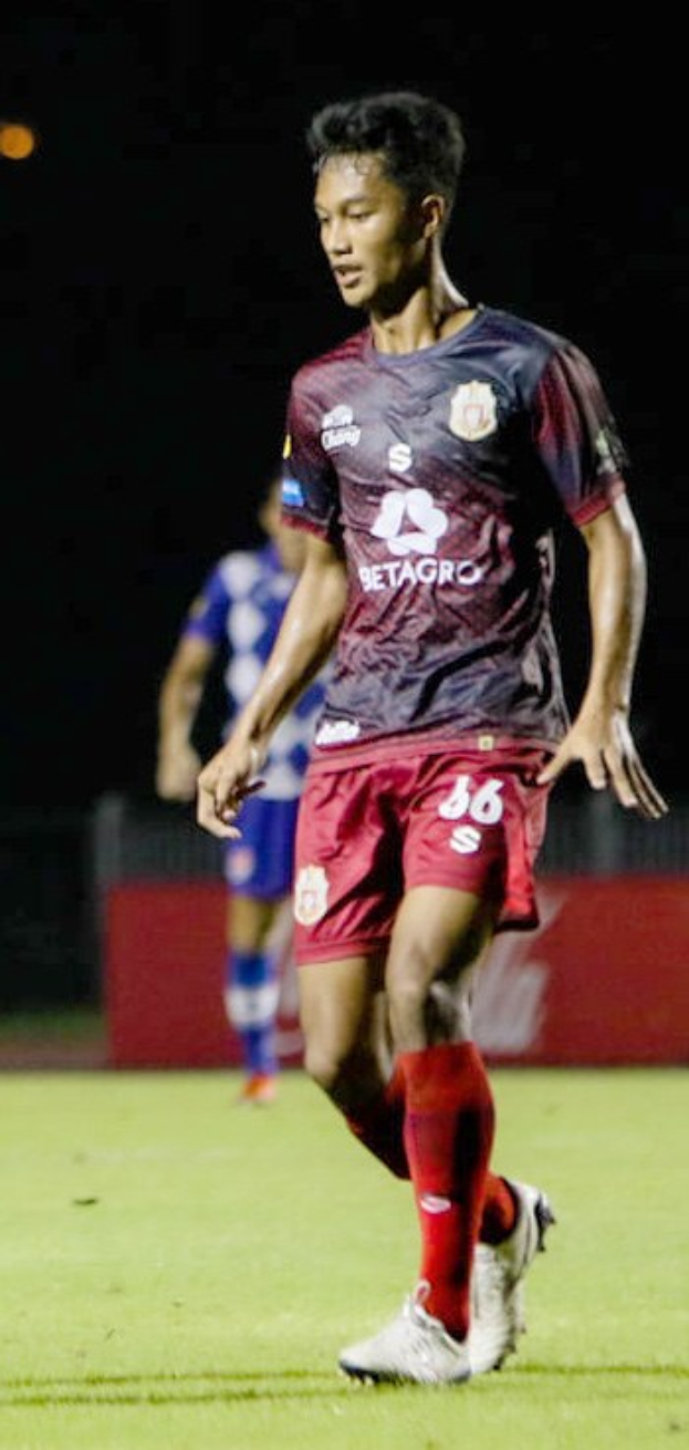 Ein Mann in einem marinefarbenen Hemd und roten Shorts, der auf einem Feld Fußball spielt, mit einer anderen Person im Hintergrund und einer Tafel mit Text.