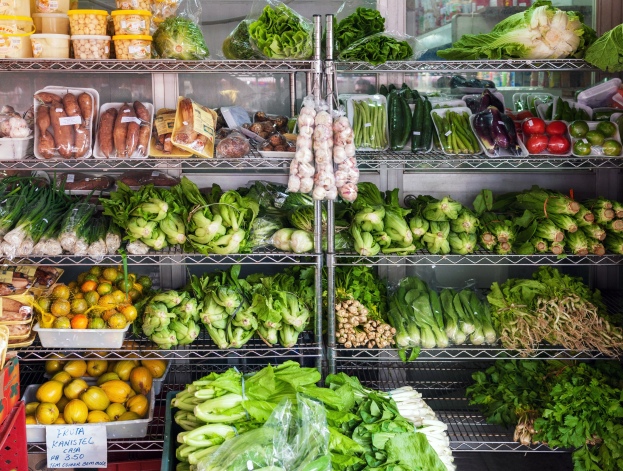 Ein Gang in einem Supermarkt mit frischem Gemüse und Obst in Plastikfolie, Kisten und einer Texttafel sowie einem Glasfenster im Hintergrund.