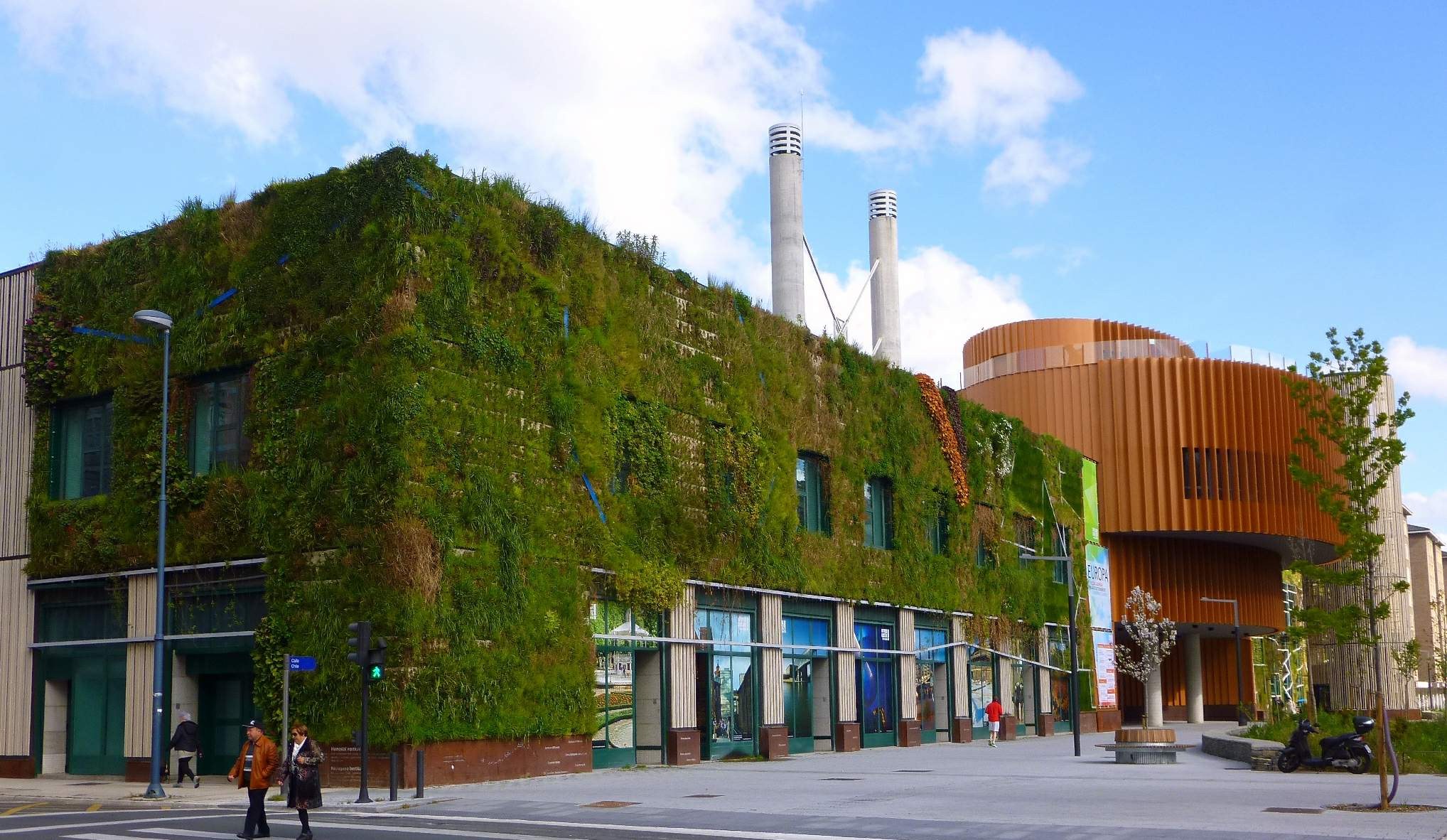 Ein großes grünwandiges Gebäude mit Straßeninfrastruktur, Fahrzeugen, Fußgängern, Bäumen und einem bewölkten Himmel, Teil eines nachhaltigen Green Building-Projekts.