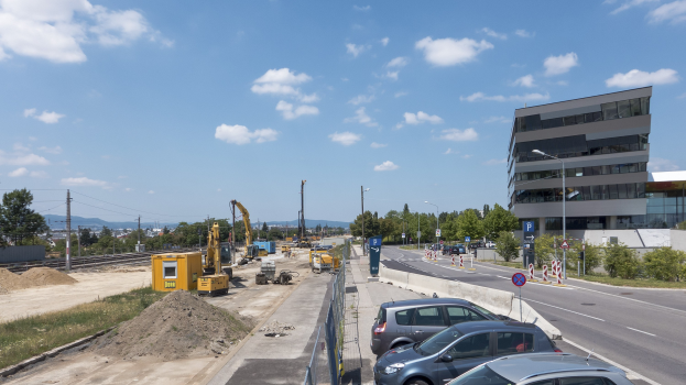 Baustelle mit parkenden Autos, Gebäuden, Straßeninfrastruktur, Vegetation, Sand, Kränen, Bahnschienen, Strommasten, Hügel und bewölktem Himmel.