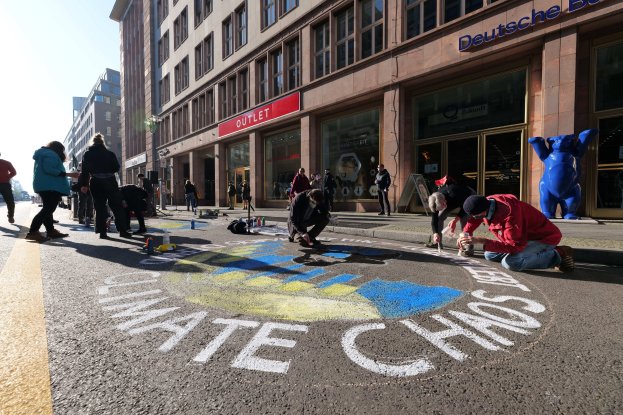 Eine Gruppe von Menschen sitzt vor einem Gebäude mit Fenstern und Namensschildern auf dem Boden, umgeben von Flaschen und anderen Gegenständen, während sie an einer Klimaschutz-Demonstration in Berlin teilnehmen, die von Bäumen und einem klaren blauen Himmel umgeben ist.