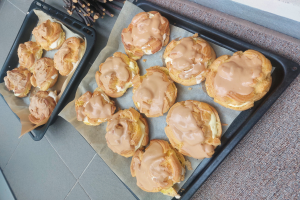 Ein Tablett voller verschiedener dänischer Kekse mit leicht goldbrauner Farbe und flaky Textur, einige mit Puderzucker bestäubt, liegt auf einer Theke neben einem offenen Fenster und wird von warmem Licht beschienen.