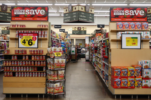 Ein Kiosk-Innenraum mit Regalen und Boden voller verschiedener Lebensmittel, Deckenleuchten, Informationsschildern und einer Person im Hintergrund.