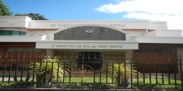 Ein Gebäude mit Fenstern, einer Tür und einem Schild mit der Aufschrift "St. Vincent Augen-, Ohren-, Nase- und Rachenklinik", umgeben von einem Metallzaun, Topfpflanzen, Gras, einem Weg und Bäumen unter einem bewölkten Himmel.
