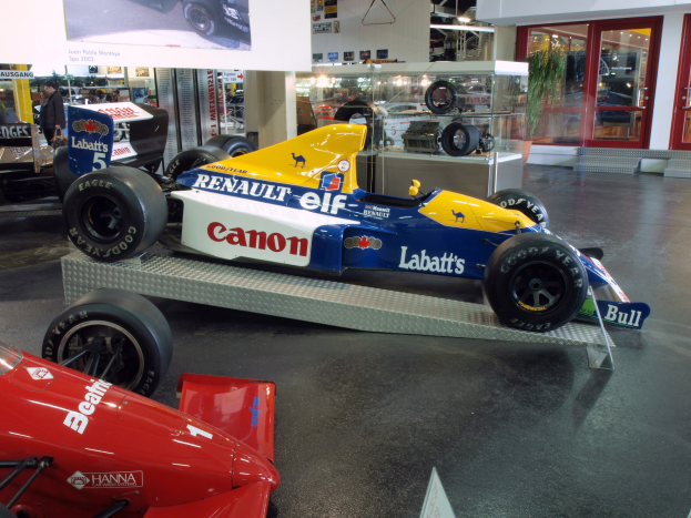 Eine Museumsausstellung zeigt verschiedene Rennwagen, darunter ein Renault-Formel-1-Auto, mit Besuchern, die beobachten, und einer Texttafel an der Wand unter beleuchteter Decke und Glastüren.