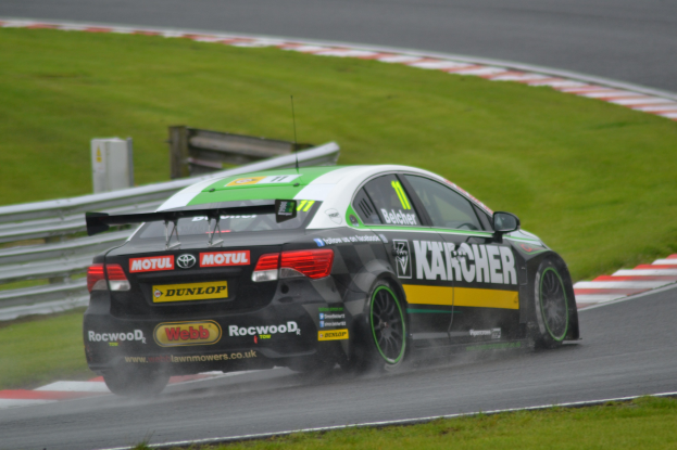 Ein Auto mit Text auf der Seite fährt auf einer regennassen Rennstrecke, die von Gras gesäumt ist, mit einer Begrenzung und einem hölzernen Gebäude im Hintergrund.