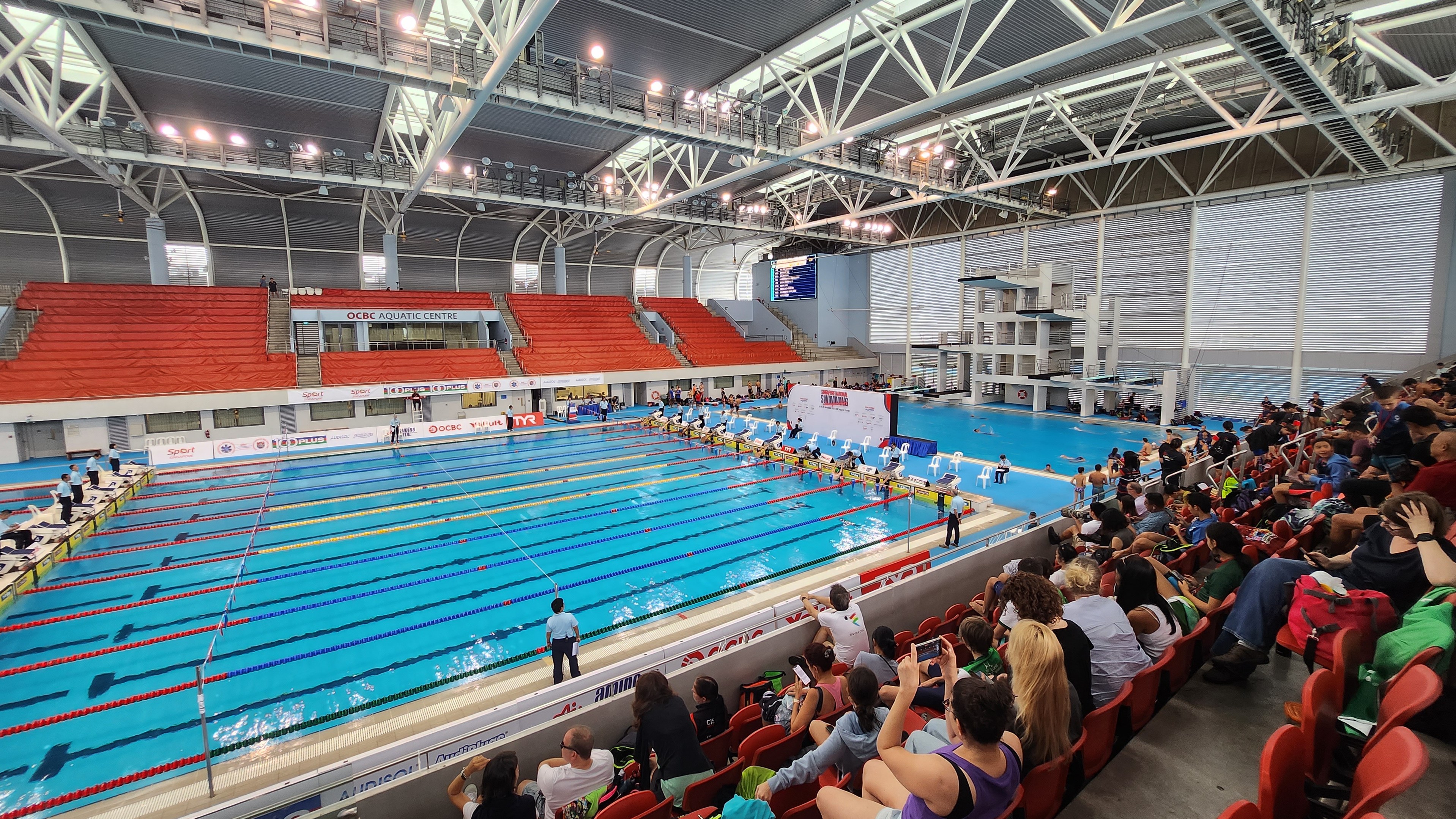 Großer Hallenbad mit vielen Menschen auf Stühlen drumherum und einigen im Wasser Stehenden, mit hoher Decke mit Lampen und Eisenstangen und markierten Tafeln im Hintergrund.