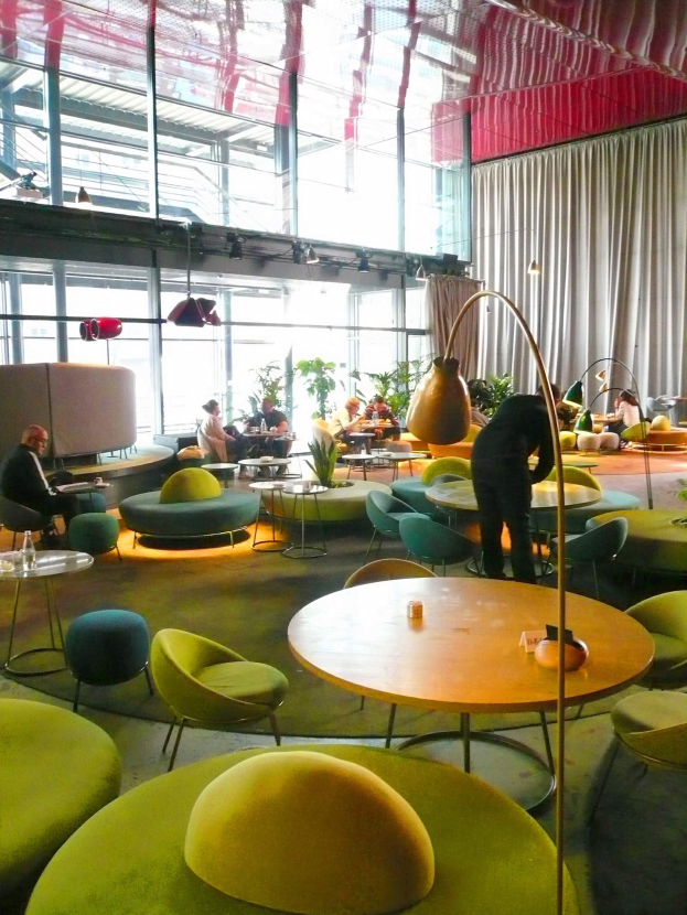 Ein geräumiger Hotellounge mit grünen Stühlen, Tischen, Sofas, stehenden Menschen, Pflanzen, einer Stehlampe, Vorhängen, Deckenleuchten und einer gemütlichen Atmosphäre.