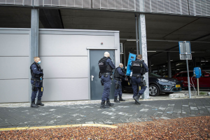Gruppe von Polizeibeamten in einer Parkanlage mit verstreuten Steinen, Fahrzeugen, Pfählen und Brettern vor einem Gebäude mit einer Tür.