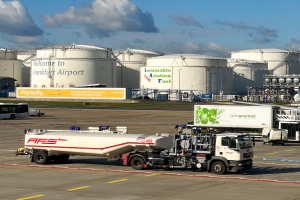 Ein großer weißer und grüner Tanklastzug parkt vor einem Gebäude am Frankfurter Flughafen, umgeben von anderen Fahrzeugen, großen weißen Tanks und einem gelben Schild im Hintergrund unter einem klaren blauen Himmel.