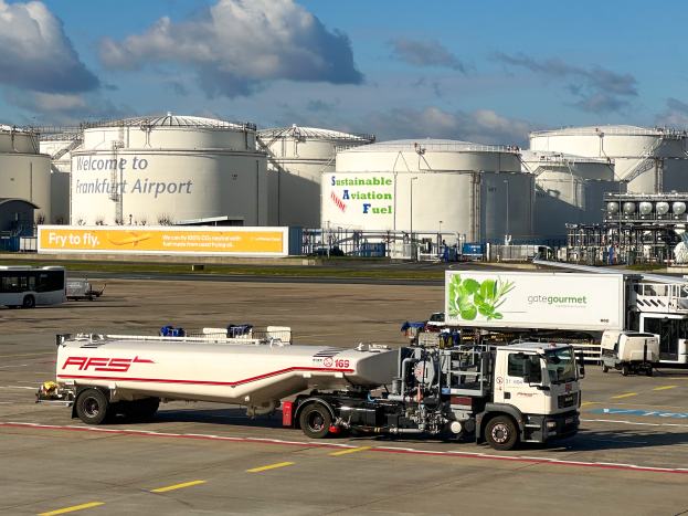 Ein großer weißer und grüner Tanklastzug parkt vor einem Gebäude am Frankfurter Flughafen, umgeben von anderen Fahrzeugen, großen weißen Tanks und einem gelben Schild im Hintergrund unter einem klaren blauen Himmel.